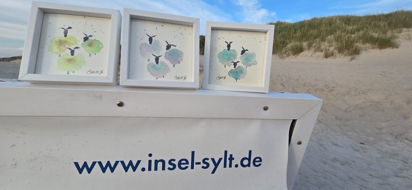 Konfettischafe Aquarell Drei gerahmte Bilder mit abstrakten, bunten Mustern auf einem Tisch am Strand.