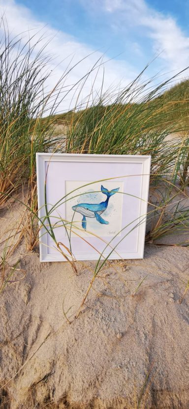 Wal Aquarell Ein blaues Fischmotiv auf einer weißen Leinwand, umgeben von Sand und Gras.
