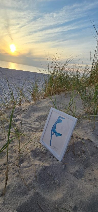Sylt Aquarell Ein Schild mit einer blauen Illustration steht in einem Sandfeld bei Sonnenuntergang.